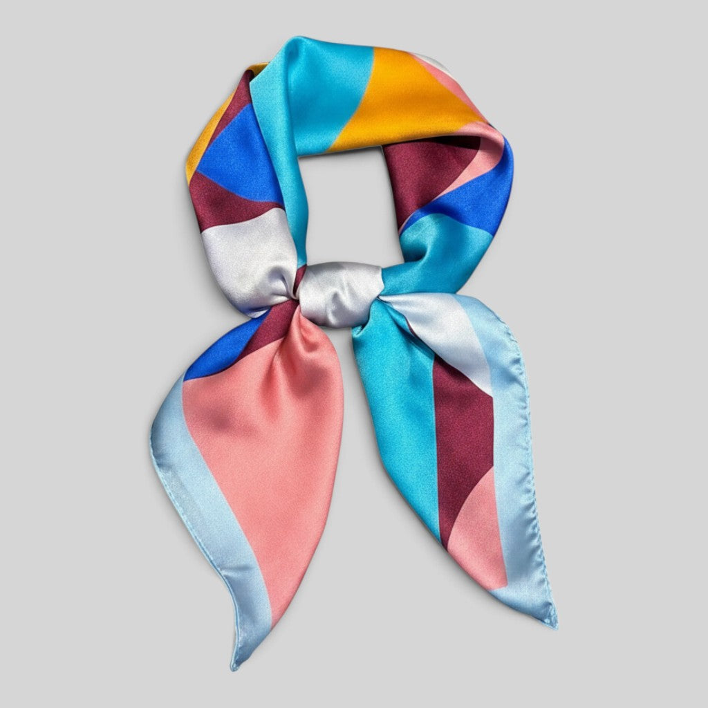 A square silk scarf with a geometric color-block pattern, tied in a bow. The pattern features shades of turquoise, dusty pink, maroon, mustard yellow, light blue, and white on a solid light gray background.