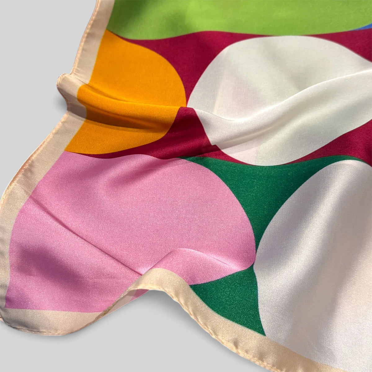 A close-up of a satin scarf with a bold abstract geometric pattern in pink, green, white, orange, and burgundy, finished with a cream border, draped on a light gray background.