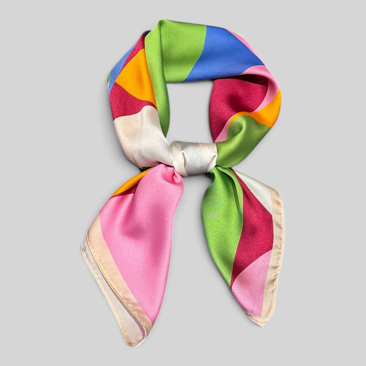 A multicolor silk scarf with a geometric color-block pattern, tied in a bow on a solid light gray background.
