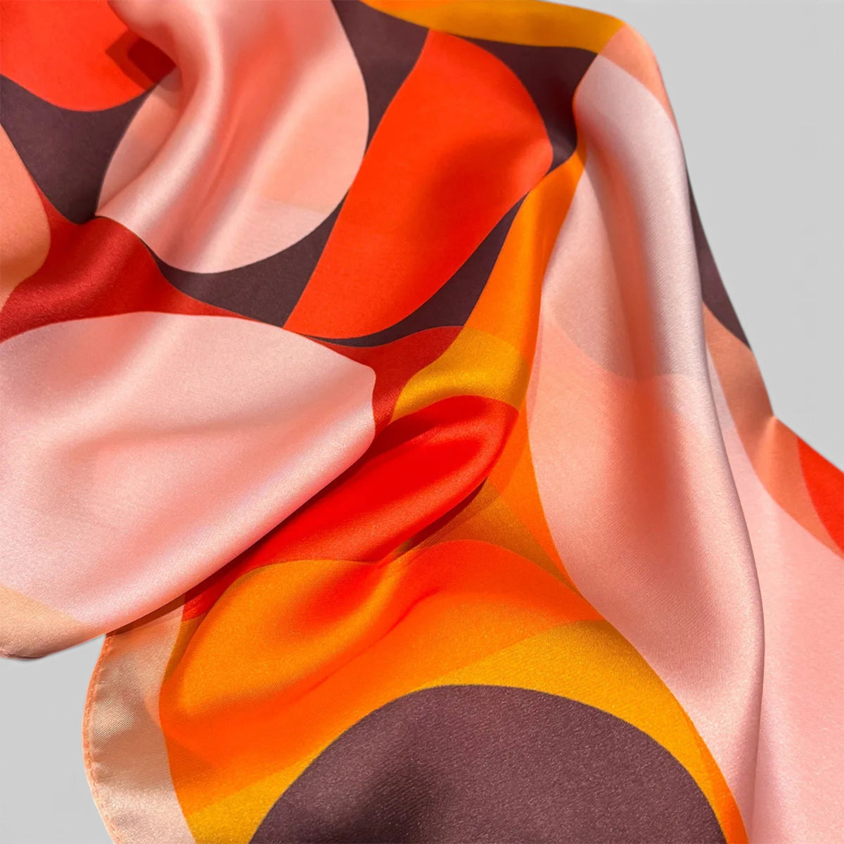 A close-up of a silky scarf with a retro abstract pattern in orange, pink, yellow, and dark brown on a light gray background.