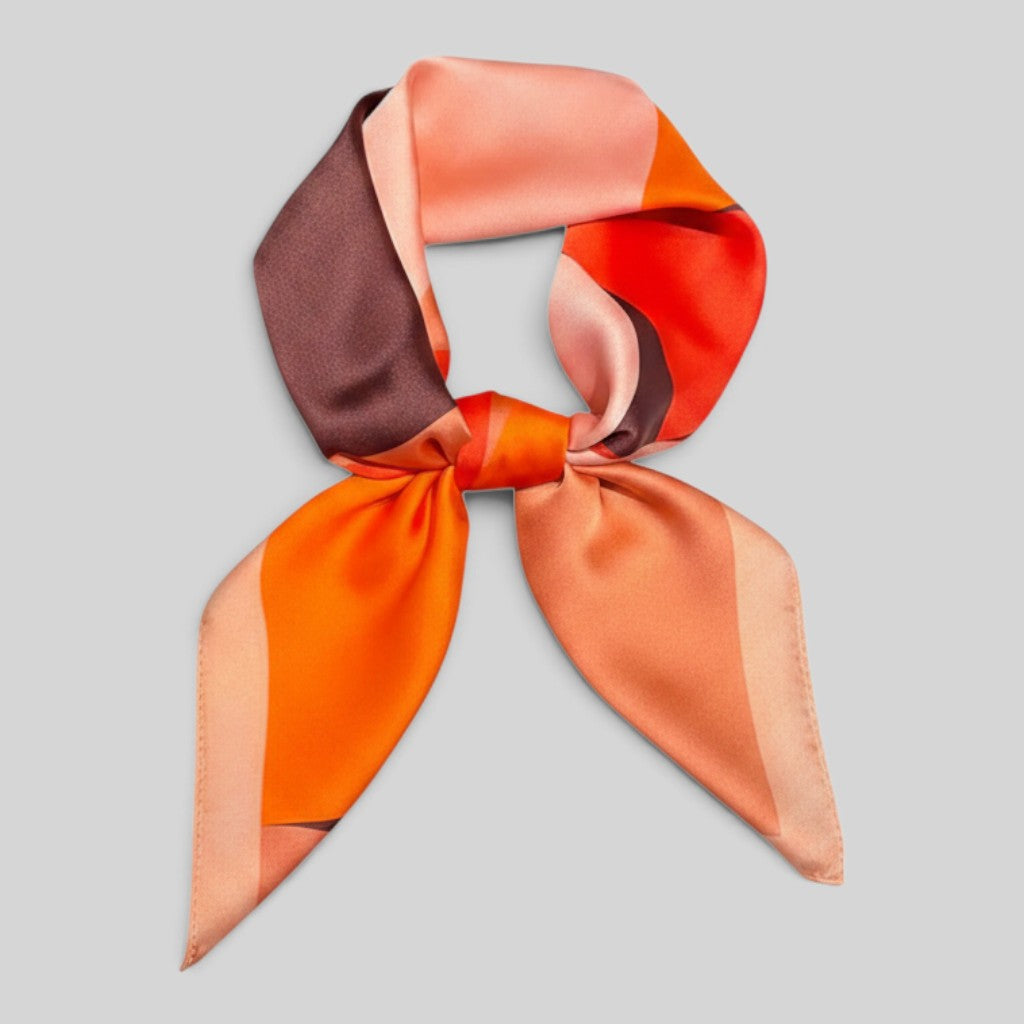 A square silk scarf with a geometric color-block pattern in orange, peach, brown, and red, tied in a bow against a solid gray background.