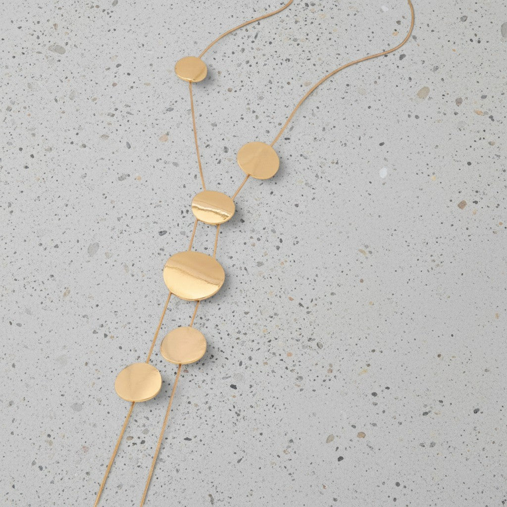 A gold disc pendant necklace with multiple circular metal charms on a fine chain, laid flat on a speckled light gray stone surface.
