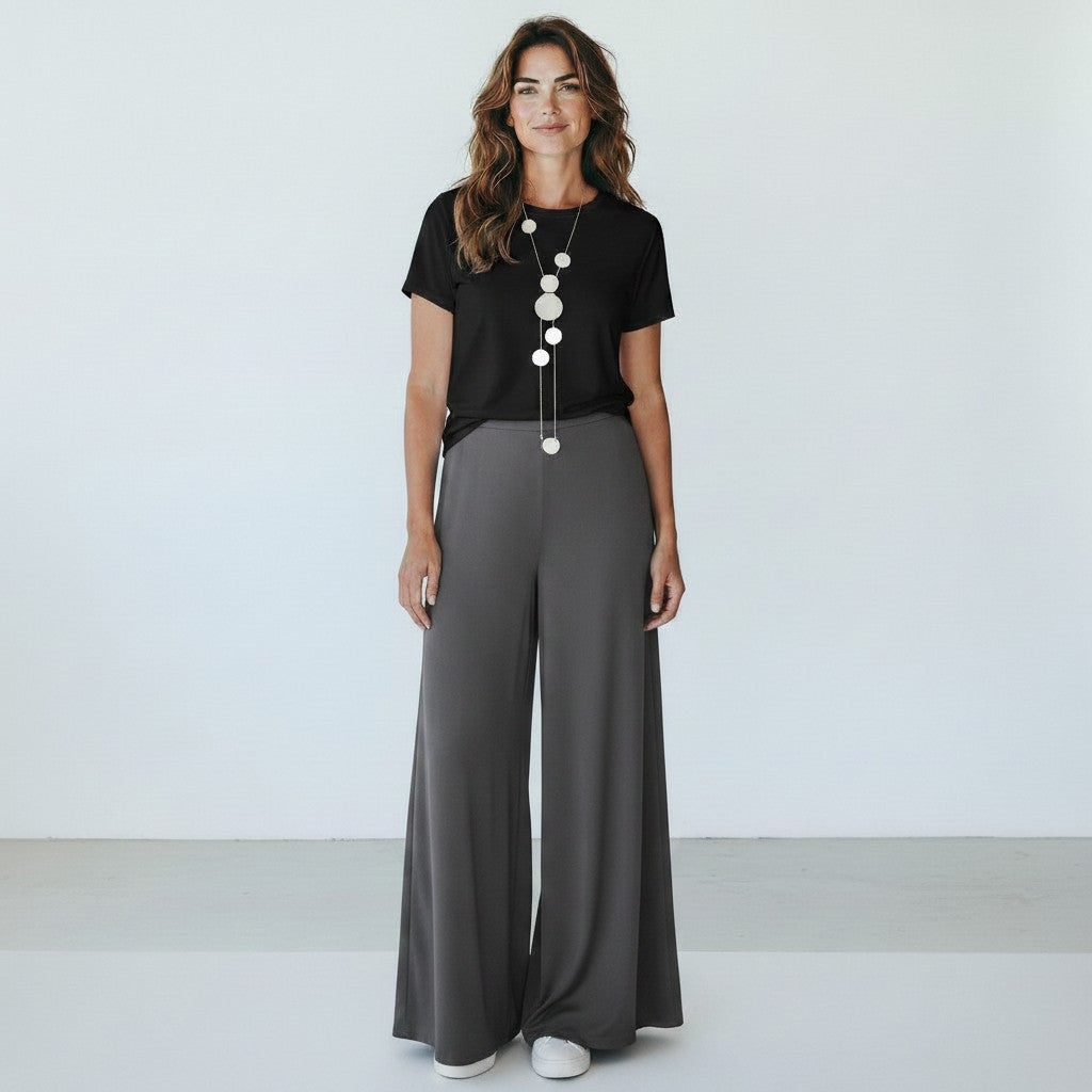 A woman stands against a plain white studio background wearing a black short-sleeve top, dark gray wide-leg pants, and a long silver pendant necklace.