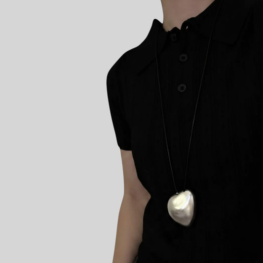 A close-up of a person wearing a black short-sleeve collared shirt and a long black cord necklace with a large, irregularly shaped white heart pendant.