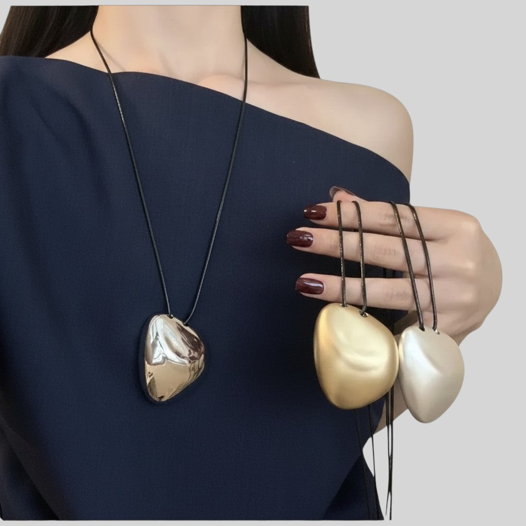 A model wearing a dark navy off-the-shoulder top displays three heart-shaped pendant necklaces. One necklace has a dark metallic, textured heart pendant on a black cord.