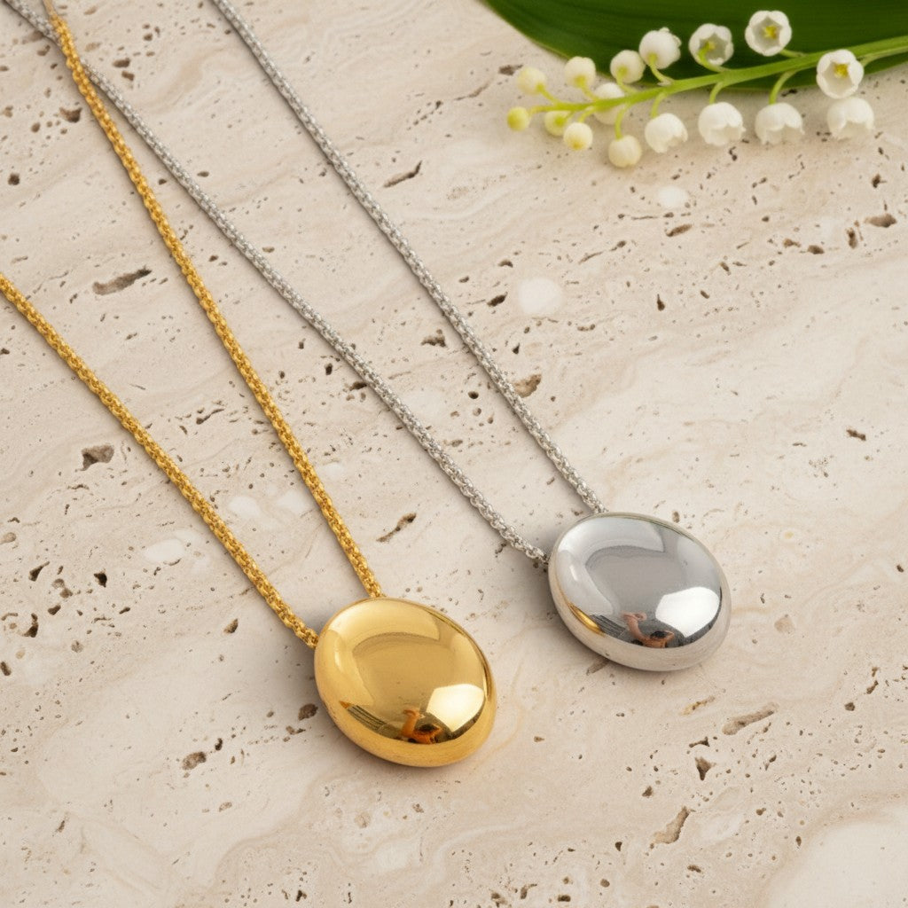Two oval pendant necklaces on textured chains, one in polished yellow gold and one in polished silver, displayed on a light beige stone surface with small white flowers in the corner.