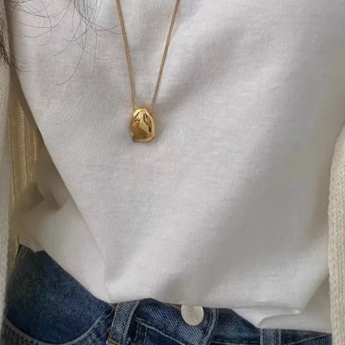 A close-up of a person wearing a gold pendant necklace with an organic, polished stone-like shape over a white top and blue jeans.