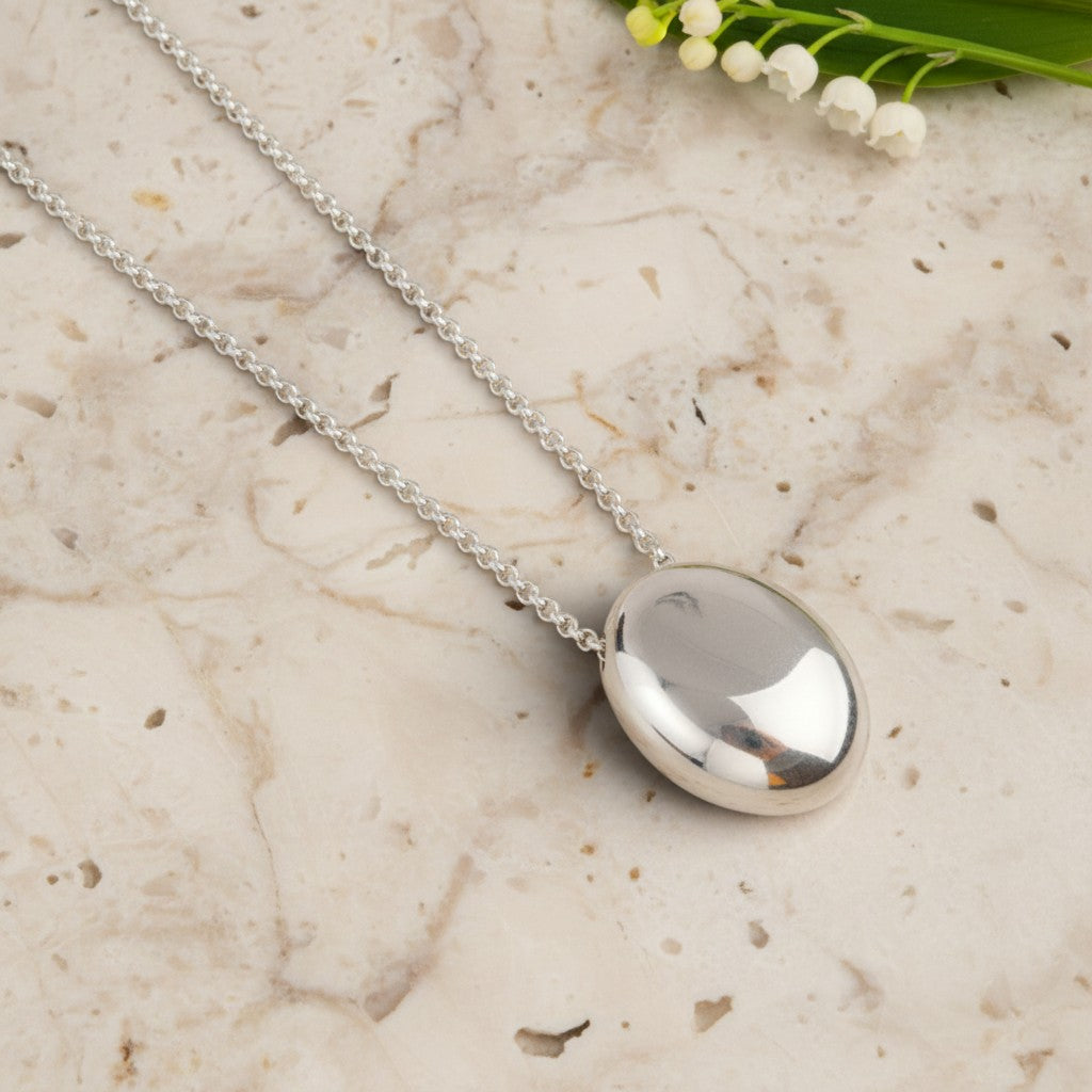 A polished silver oval pendant necklace on a delicate silver chain, displayed on a light beige marble surface with small white flowers in the corner.