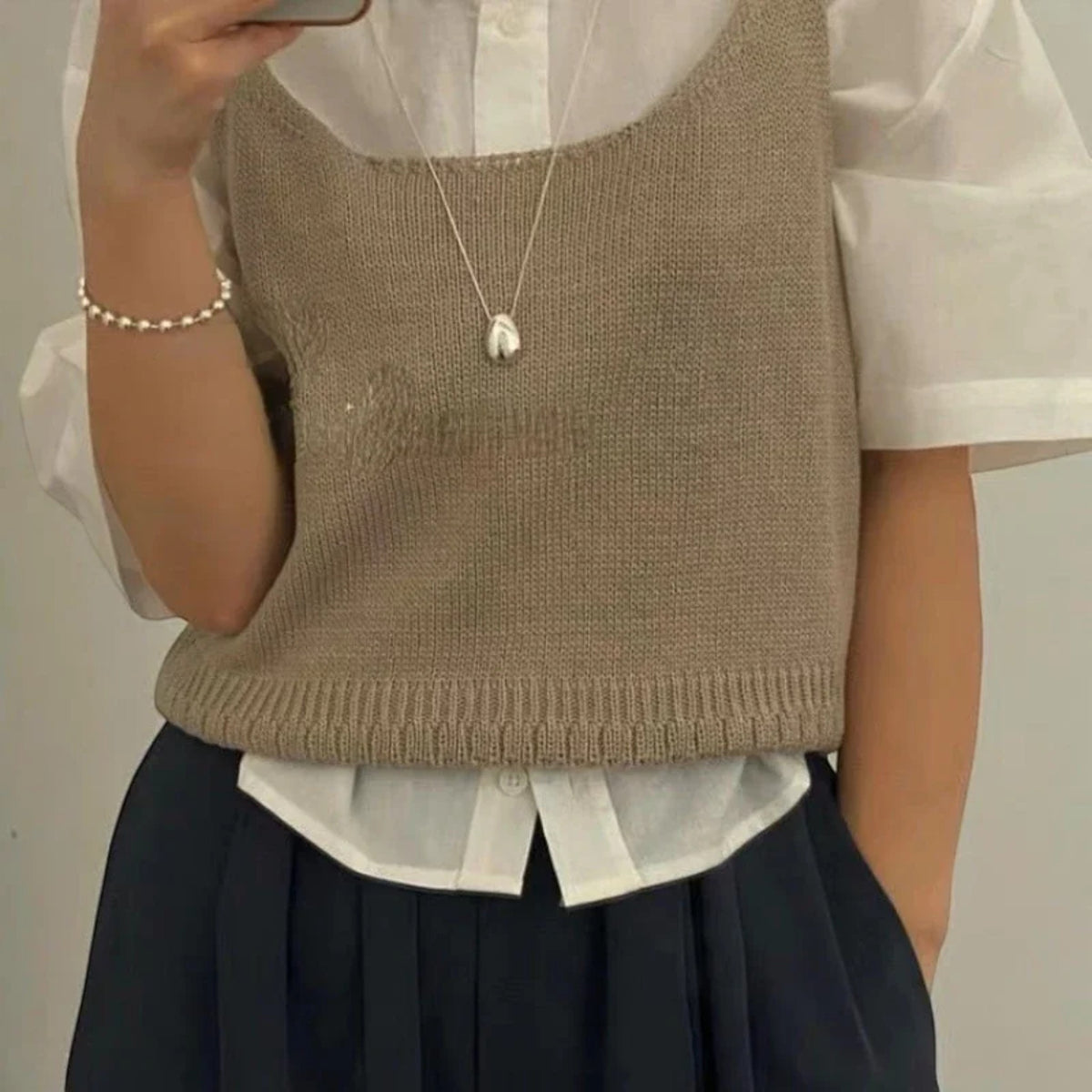 Person wearing a beige knitted vest over a white shirt with a pearl necklace.