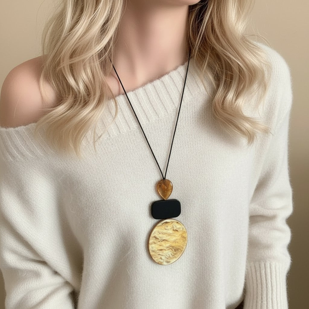 A close-up of a woman wearing a modern multi-layered pendant necklace. The necklace features a black cord, a small gold heart, a black rectangular block, and a large textured gold oval pendant.