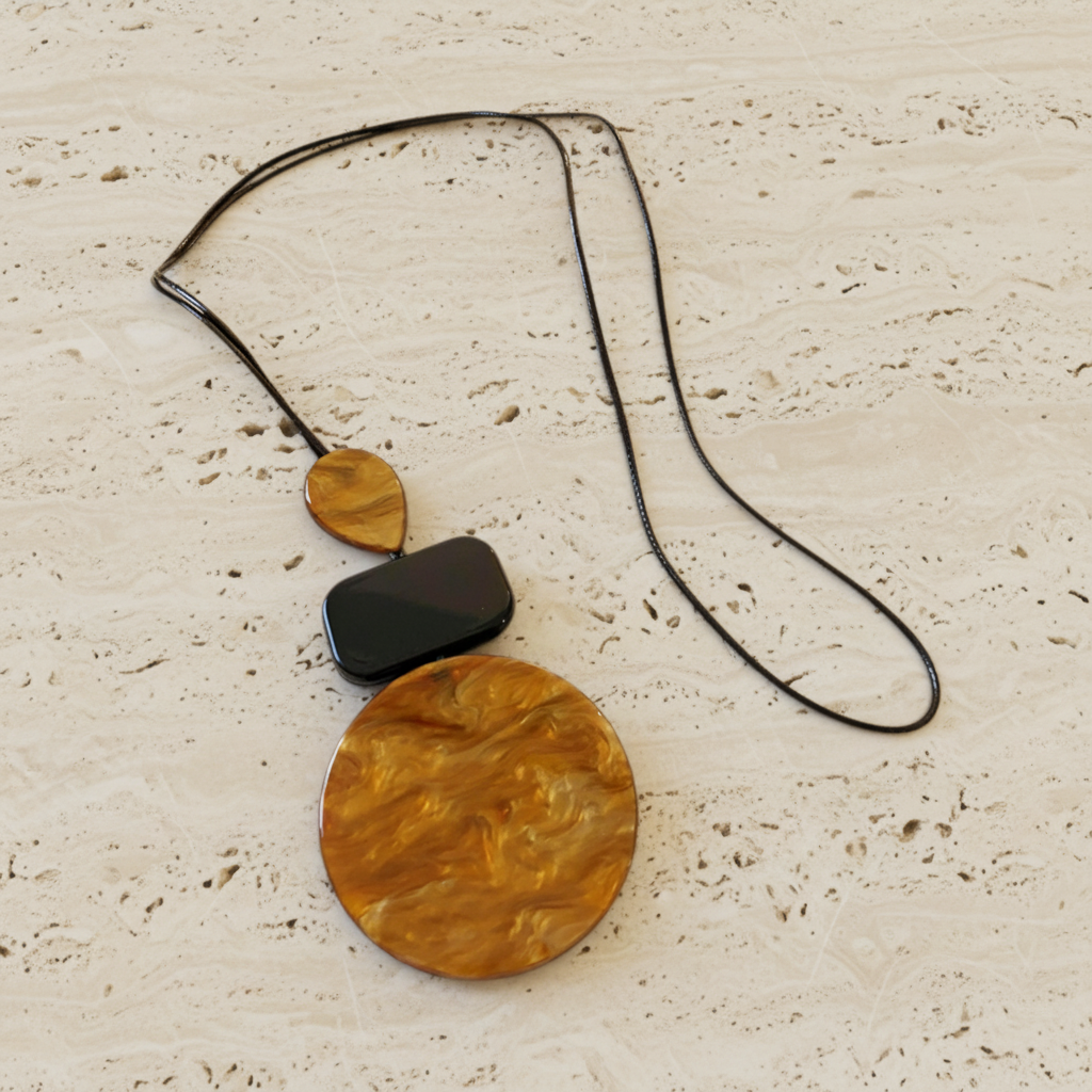 A modern necklace featuring a large circular and a smaller teardrop-shaped amber marbled pendant, connected by a black rectangular link and a thin black chain, displayed on a textured beige stone surface.