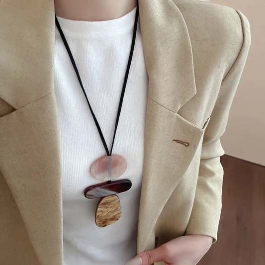 A close-up of a person wearing a cream-colored linen blazer over a white ribbed knit top, featuring a black cord necklace with stacked stone and wood pendants.