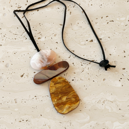 A black cord necklace with three polished stone pendants in white, brown, and amber-yellow marbled shapes, arranged on a light beige textured surface.