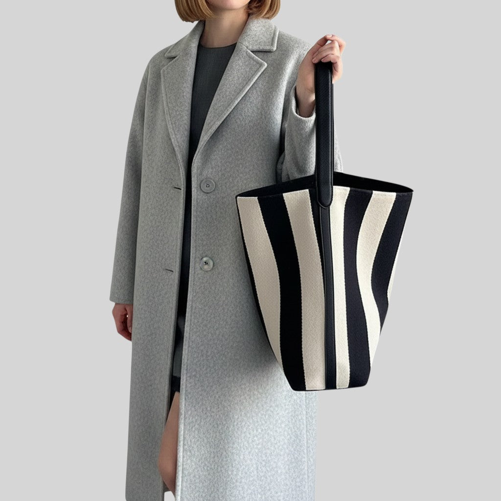 A model wearing a light gray overcoat holds a large black and cream striped canvas tote bag against a solid gray background.