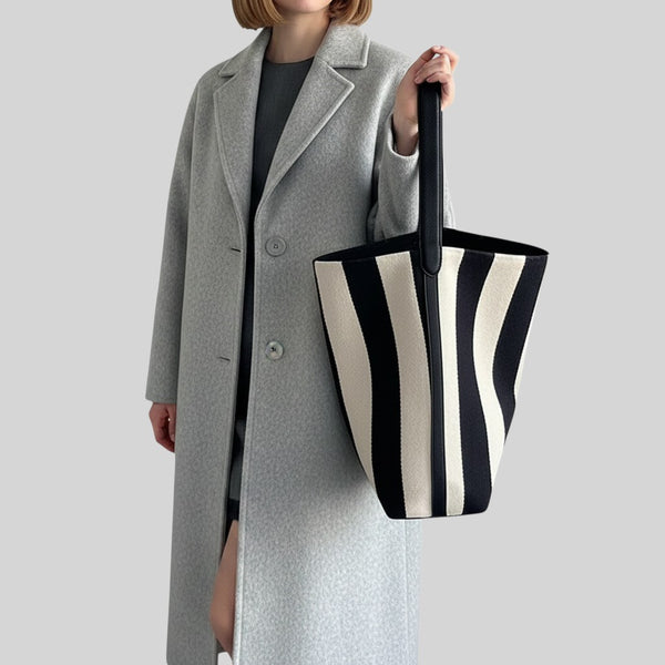 A model wearing a light gray overcoat holds a large black and cream striped canvas tote bag against a solid gray background.