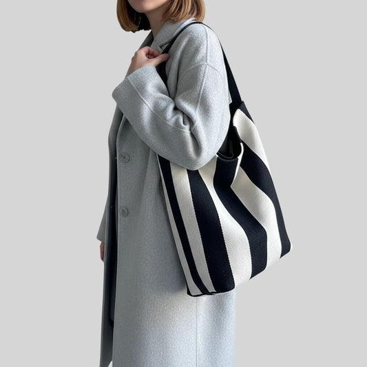 A woman wearing a light gray coat carries a large black and white striped knit tote bag over her shoulder against a plain light gray background.